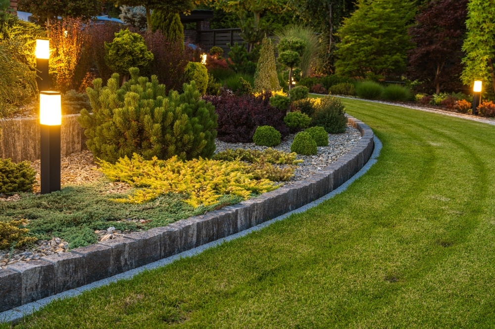 Jardin paysager avec bordure en pierre, pelouse tondue, arbustes variés et éclairage extérieur chaleureux.