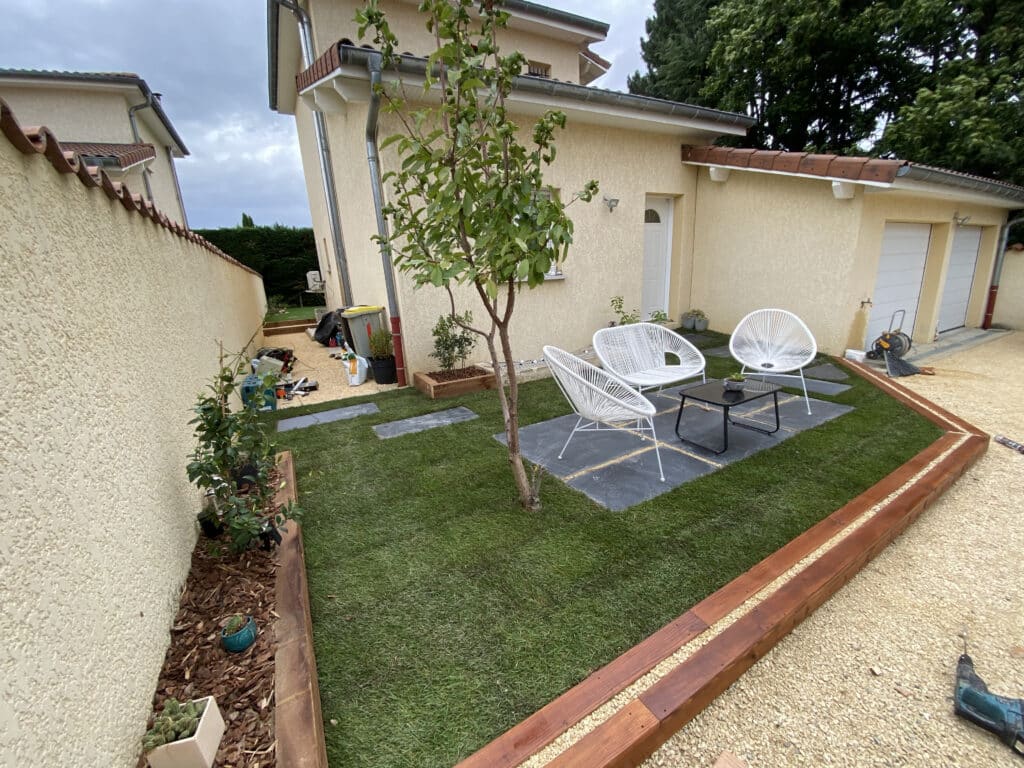 Aménagement de jardin moderne avec terrasse et pelouse Petit jardin moderne avec chaises blanches, terrasse en ardoise, pelouse verte et bordure en bois devant une maison.