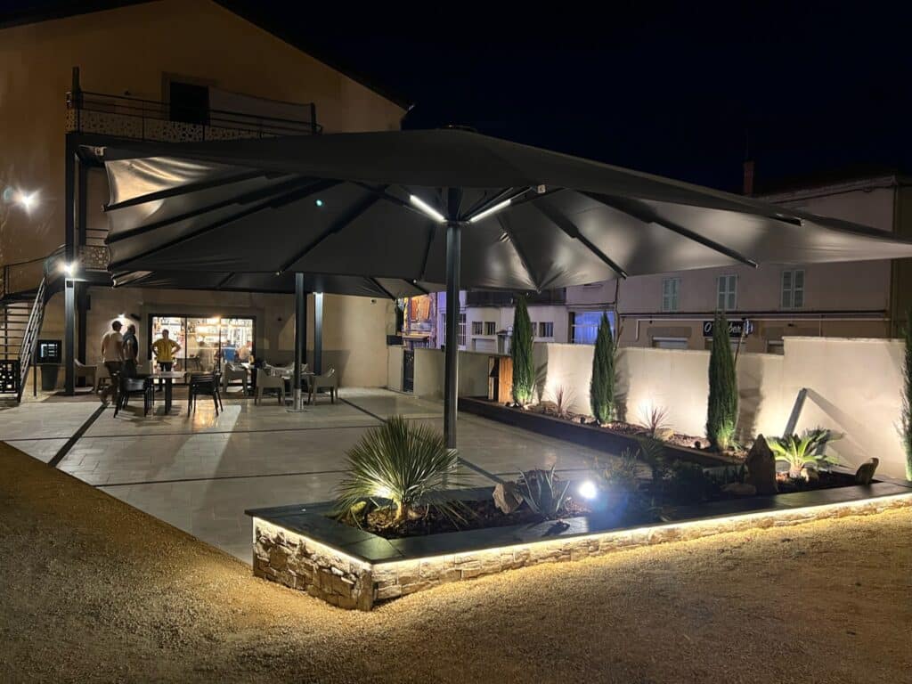 Parasol Géant LED pour Terrasse de Restaurant Moderne Terrasse nocturne moderne avec grand parasol gris éclairé, jardinières en pierre illuminées et mobilier de jardin.