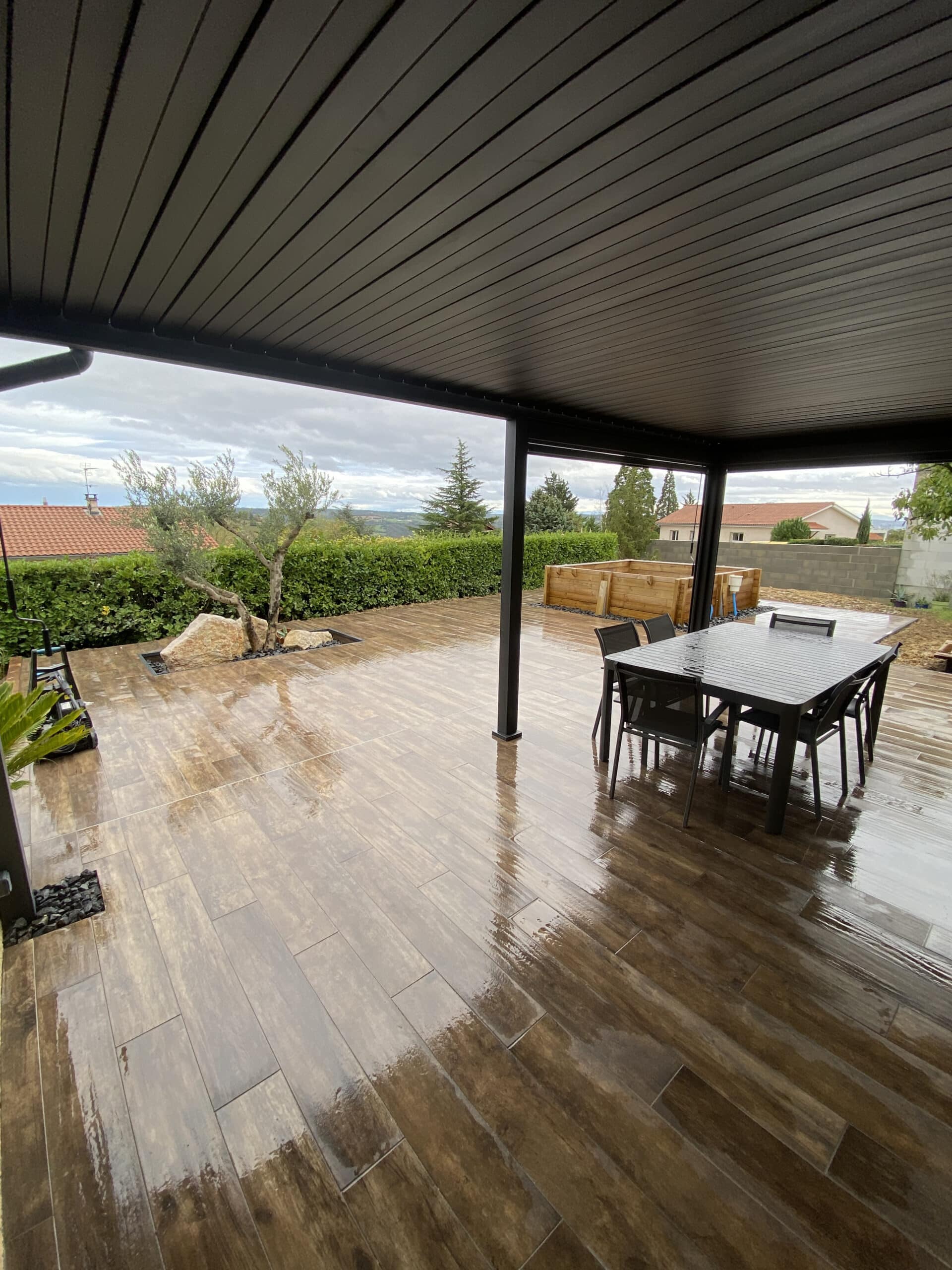Terrasse moderne avec pergola noire, carrelage aspect bois et mobilier de jardin par temps de pluie.